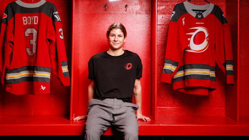 Zoe Boyd, a PWHL defenceman for the Ottawa Charge, seated with a smile, framed by team jerseys hanging behind her
