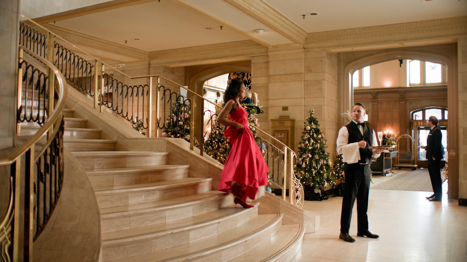 Sarah walks down a sweeping staircase in a red gown. 