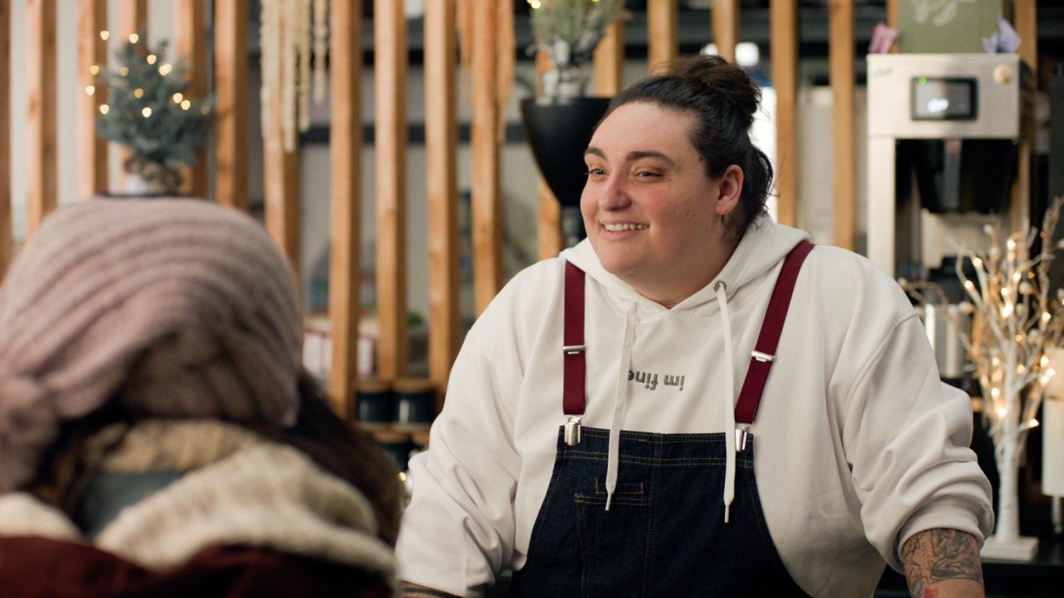 Jamie Fine as The Barista smiling, wearing an apron. 
