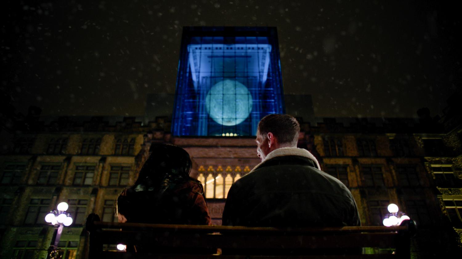 Two people sit on a bench outside, looking up at a moon replica hanging in a museum. 