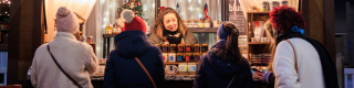 A market stand decorated with Christmas lights with several shoppers perusing. 