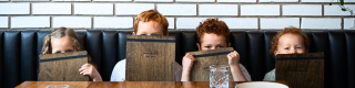 Four children sit in a restaurant booth, peeking out from behind wooden menus.